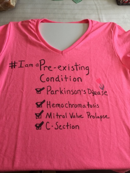Image Description: a pink t-shirt with the words "I am a pre-existing condition" and a list of my mother's illnesses, Parkinson's Disease, hemochromatosis, Mitral Valve Prolapse, and C-section